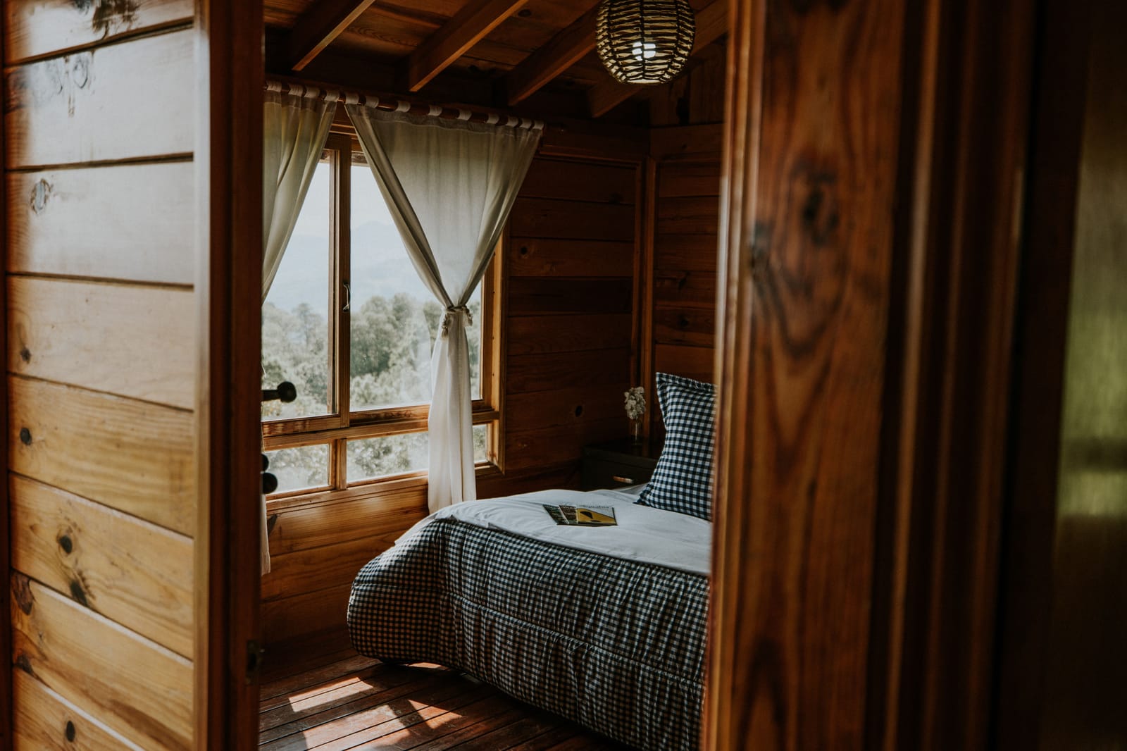 Cozy wood-paneled cabin bedroom with a gingham bed and a window looking out over trees and mountains